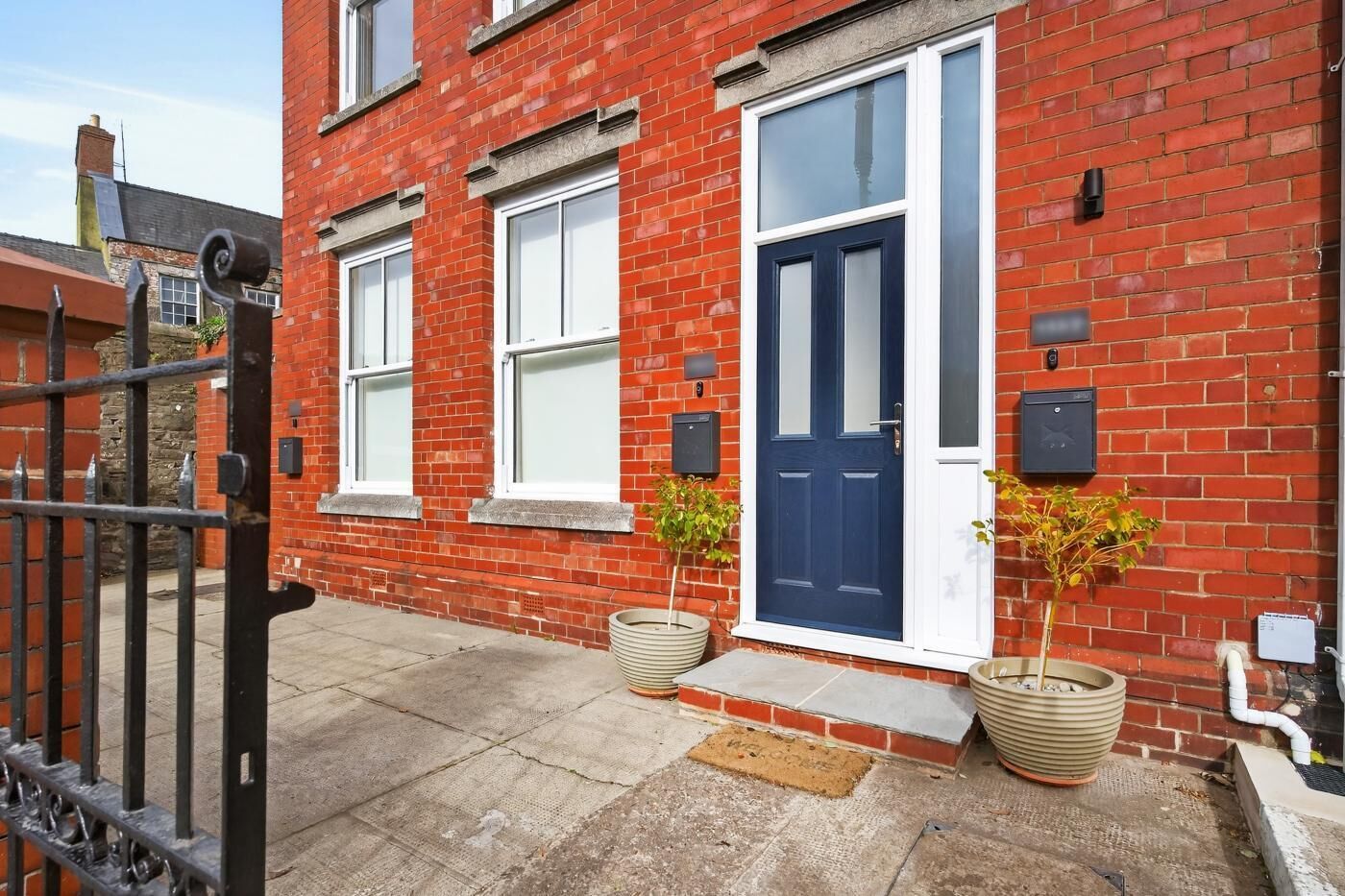 A red brick house with a blue door, two windows, a gated entrance, and two potted plants.