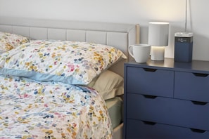 En pænt redt seng med farverigt sengetøj, et natbord med lampe og digitalt ur, og en blå kommode med skuffer.