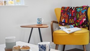 Ein gemütlicher Sitzbereich mit einem gelben Sessel, einem kleinen Holztisch, einer blauen Tasse, einer weißen Kerze und einem Teller mit Muffins.