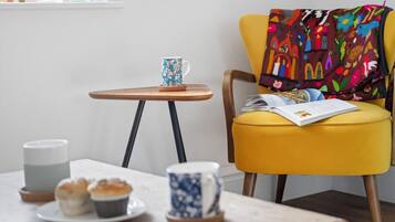 A cozy seating area with a yellow chair, a small wooden table with a blue mug, a white candle, and a plate with muffins.
