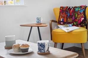 A cozy seating area with a yellow chair, a small wooden table with a blue mug, a white candle, and a plate with muffins.