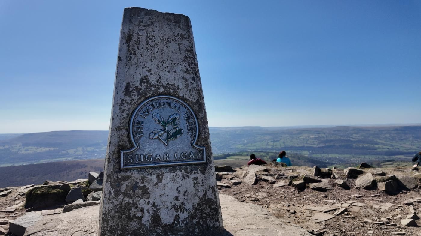 Un senyal rovellat amb la inscripció 'National Trust The Sugar Loaf' damunt un turó rocós amb vista a les colines llunyanes.