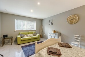 A bedroom with a bed, a green sofa, a white chair, a bedside table, a clock, and a window with blinds.