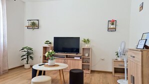 Un salon moderne avec un téléviseur à écran plat, des meubles en bois, une petite table ronde agrémentée d’une plante, un pouf noir et un ventilateur blanc.