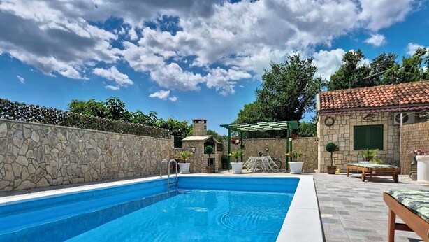 Une piscine bordée d’un mur en pierre, une pergola et un bâtiment en pierre en arrière-plan.