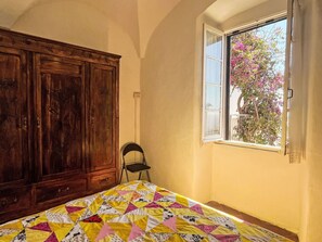 Une pièce avec un lit, une armoire en bois, une fenêtre donnant sur des fleurs roses et une chaise.