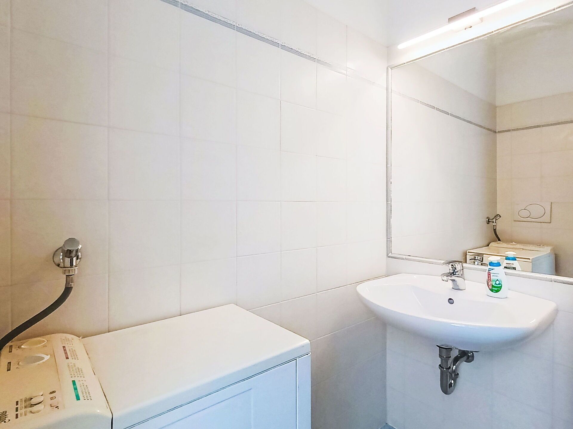 A bathroom with white tiles, a sink, a mirror, and a towel dispenser.