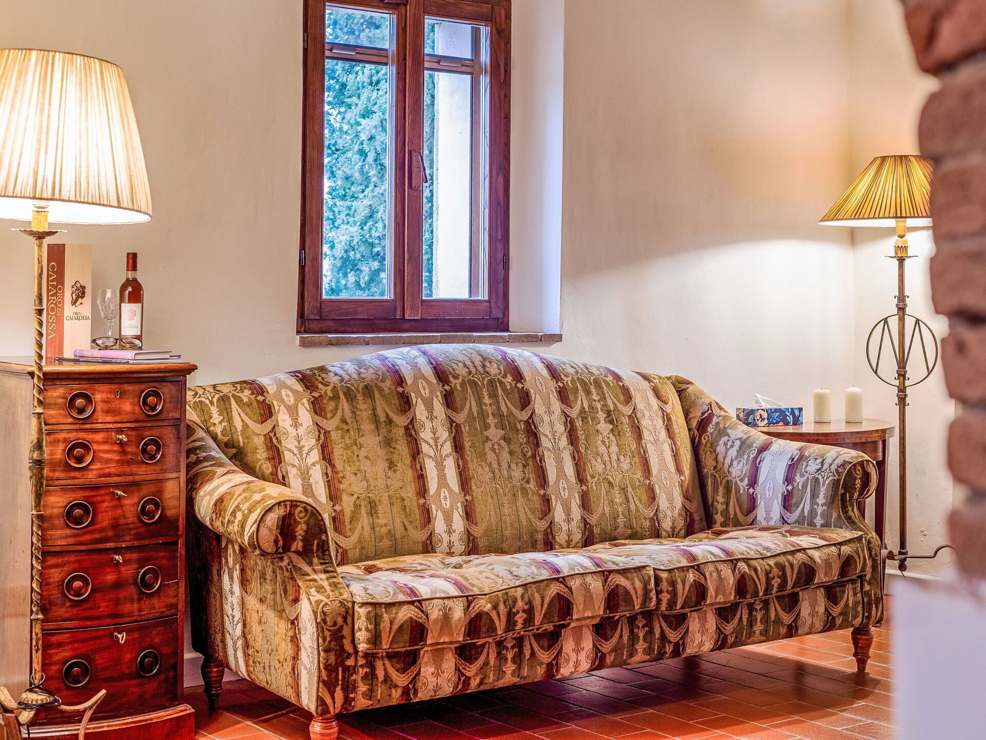Un canapé ancien recouvert d’un tissu à motifs, une commode en bois et une lampe sur pied.