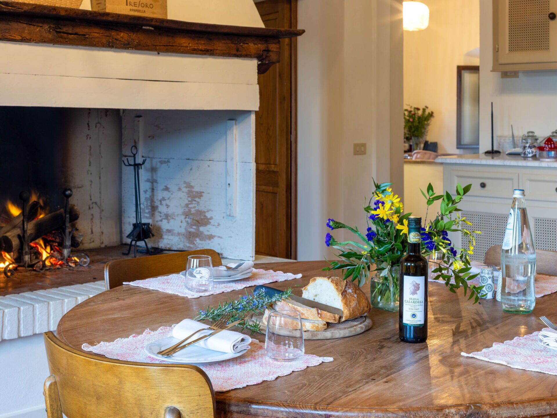 Una mesa de comedor servida con platos, cubiertos, una hogaza de pan, una botella de aceite de oliva y un florero con flores.