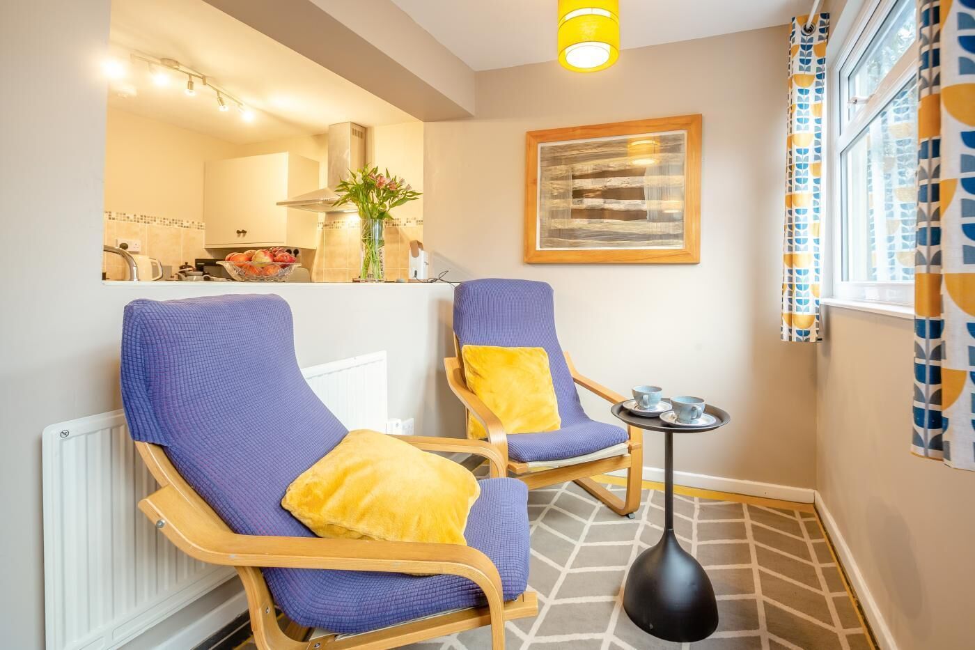A small room with two blue armchairs, a yellow cushion, a black floor lamp, a framed abstract painting, and a window with patterned curtains.
