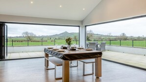 A spacious room with a large table set for dining, a bench, and a view of a green landscape through large windows. - 3 Bed in West Pennard (oc-b34974) (Coxbridge)