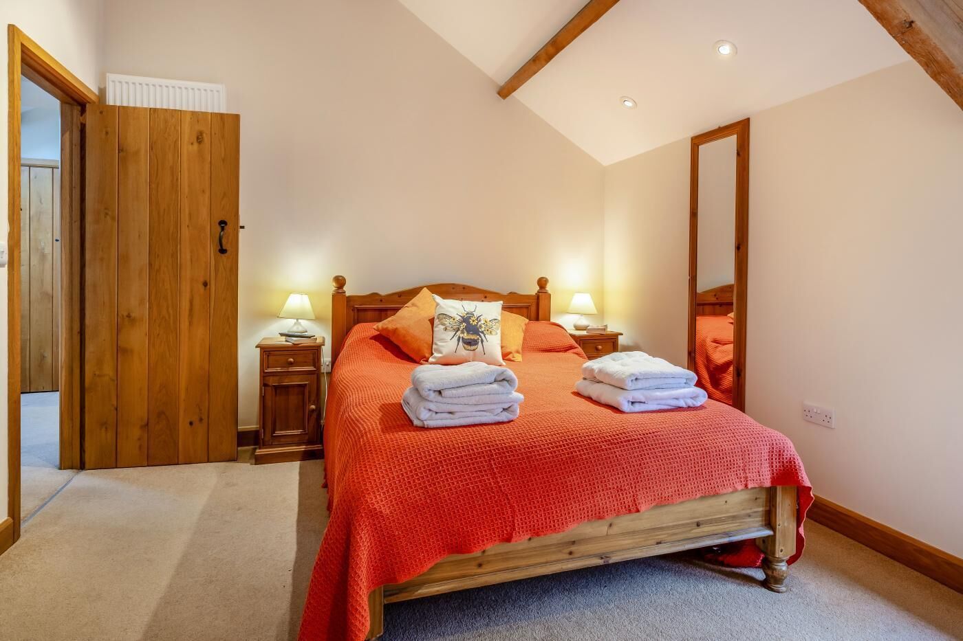 A bedroom with a wooden bed, red bedding, two bedside tables with lamps, a mirror, and a door with a radiator above it.