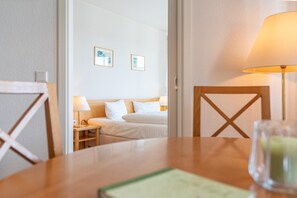 A hotel room with a wooden table, a lamp, a bed with white linens, and two framed pictures on the wall. - Ferienwohnung in Sellin mit Balkon, Hund (Sellin)