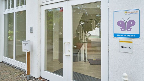 A white building with a glass door, a butterfly logo sign, and a number sign. - Ferienwohnung in Sellin mit Balkon, Hund (Sellin)