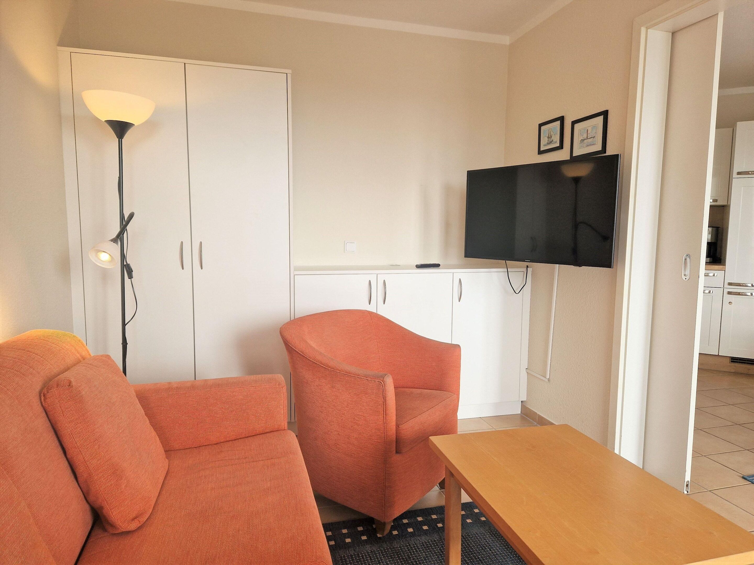 Un espace de vie compact comprenant un canapé, un fauteuil, une table en bois, un téléviseur fixé au mur et, au fond, une petite cuisine.