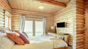 A log cabin bedroom with a bed, pillows, a TV, and a view of the ocean. - 3 Bed in Ravensworth (oc-s34987) (Ravensworth)