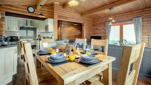 A wooden cabin interior with a dining table set for breakfast, kitchen area, and a view of the sea. - 3 Bed in Ravensworth (oc-s34987) (Ravensworth)