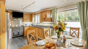 A modern kitchen with wooden cabinets, stainless steel appliances, and a dining area with a table set for two. - 3 Bed in Polruan (oc-l35010) (Fowey)