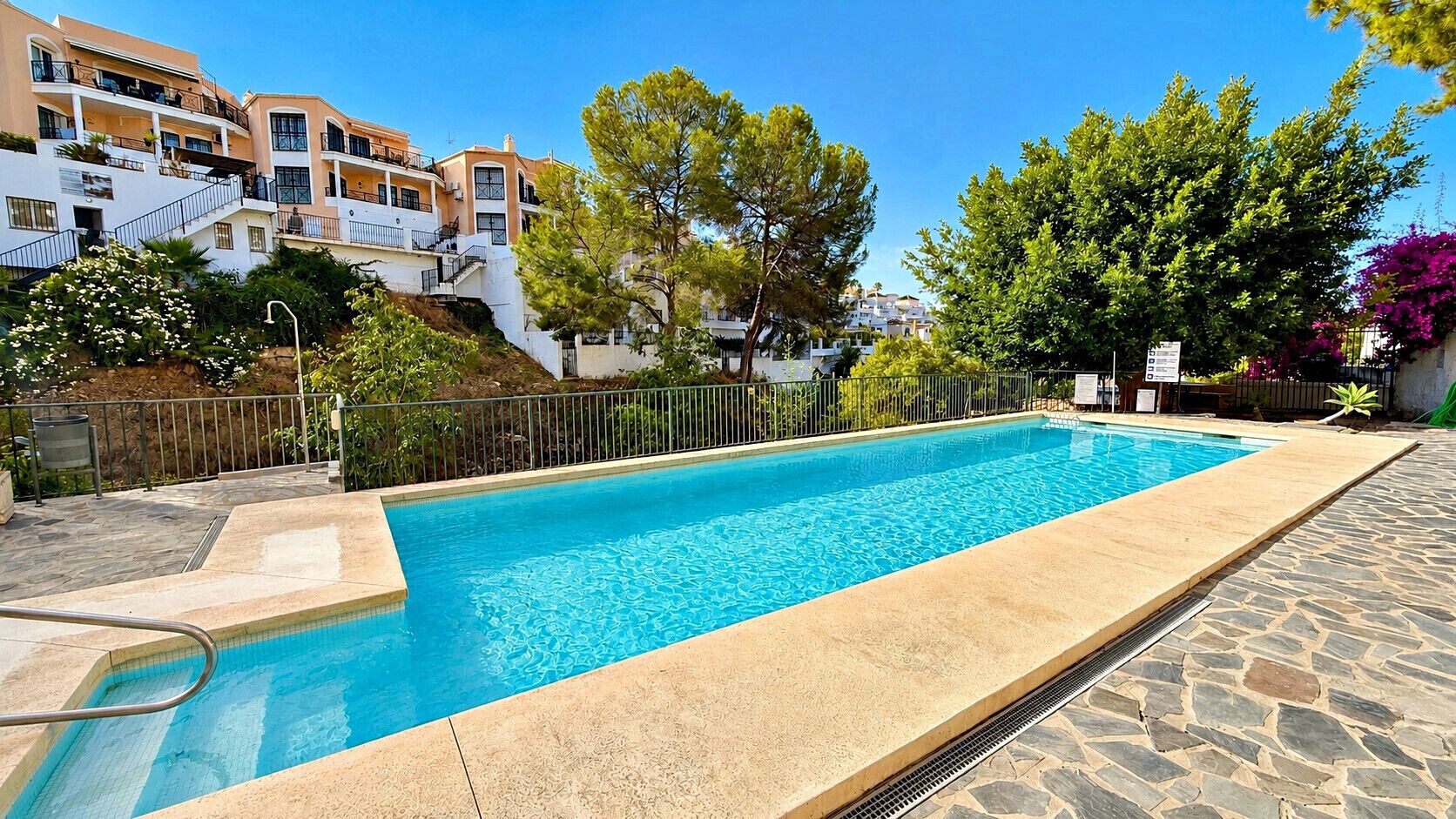 Une piscine rectangulaire avec une plage en pierre, entourée d’une clôture en métal et située à côté d’un bâtiment doté de balcons.