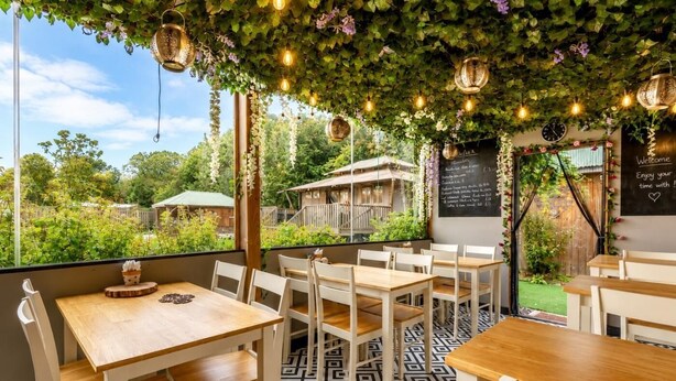 An outdoor seating area with tables and chairs, a chalkboard menu, and a view of greenery.