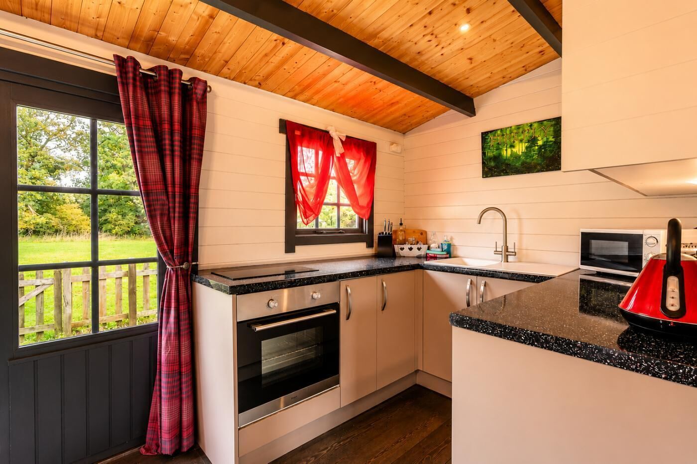 Une cuisine avec un plafond en bois, une fenêtre avec des rideaux rouges, un four à micro-ondes, une bouilloire et un évier.