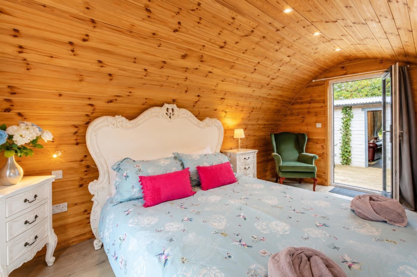 Une chambre avec un plafond en bois, un lit recouvert d’une literie à motifs floraux, une chaise verte et une table de chevet avec une lampe.