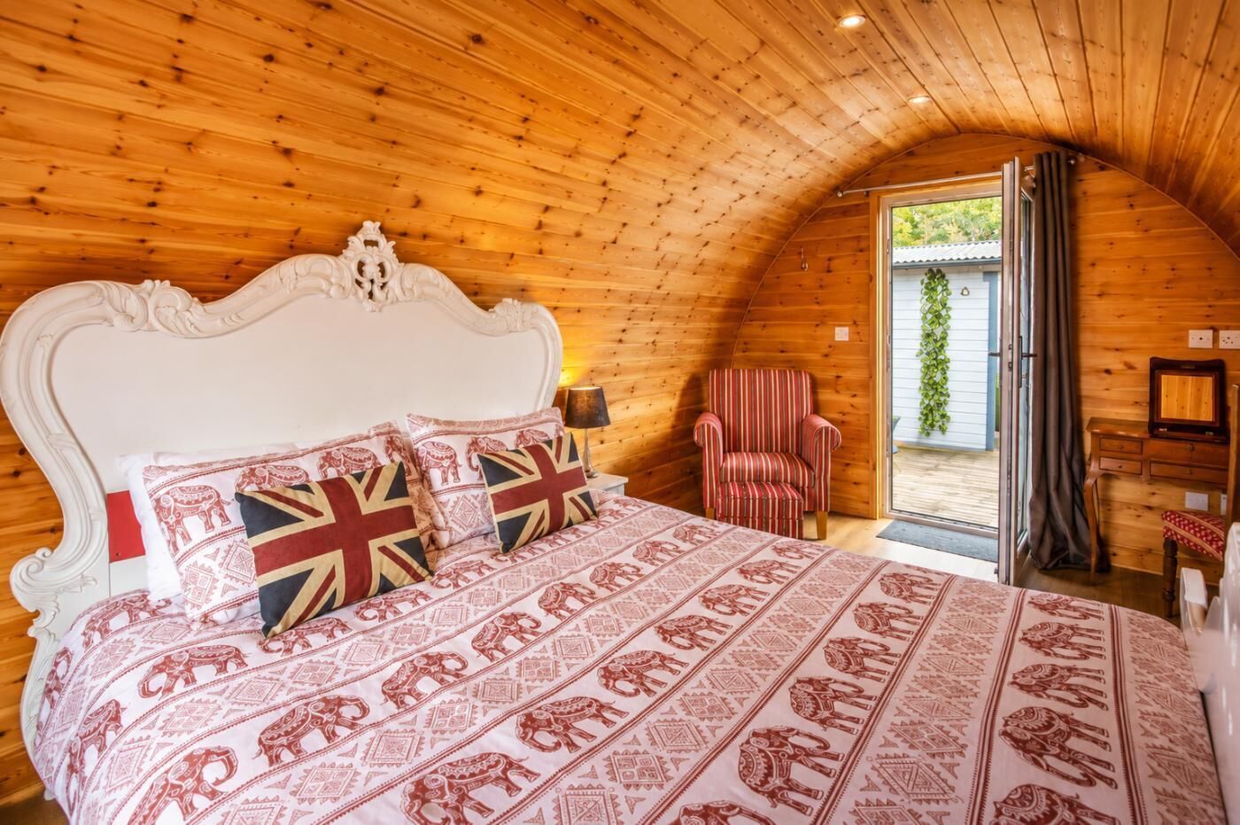 Une chambre avec un plafond en bois, une tête de lit blanche ornée, une couvre-lit à motifs, un fauteuil à rayures et une vue sur l’extérieur, visible à travers une porte ouverte.