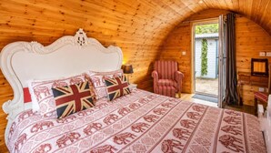 Ein Schlafzimmer mit Holzbalkendecke, einem weißen verzierten Kopfteil, einer gemusterten Bettdecke, einem gestreiften Sessel und Blick durch eine geöffnete Tür.