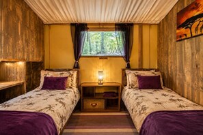 Ein Zimmer mit zwei Betten, einem Fenster mit Vorhängen, einem Nachttisch mit Lampe und einem Bild an der Wand.