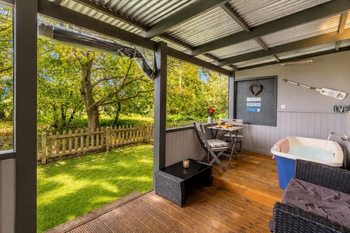 Un espace extérieur abrité, équipé d’une table et de chaises, d’une baignoire et offrant une vue sur un jardin verdoyant.
