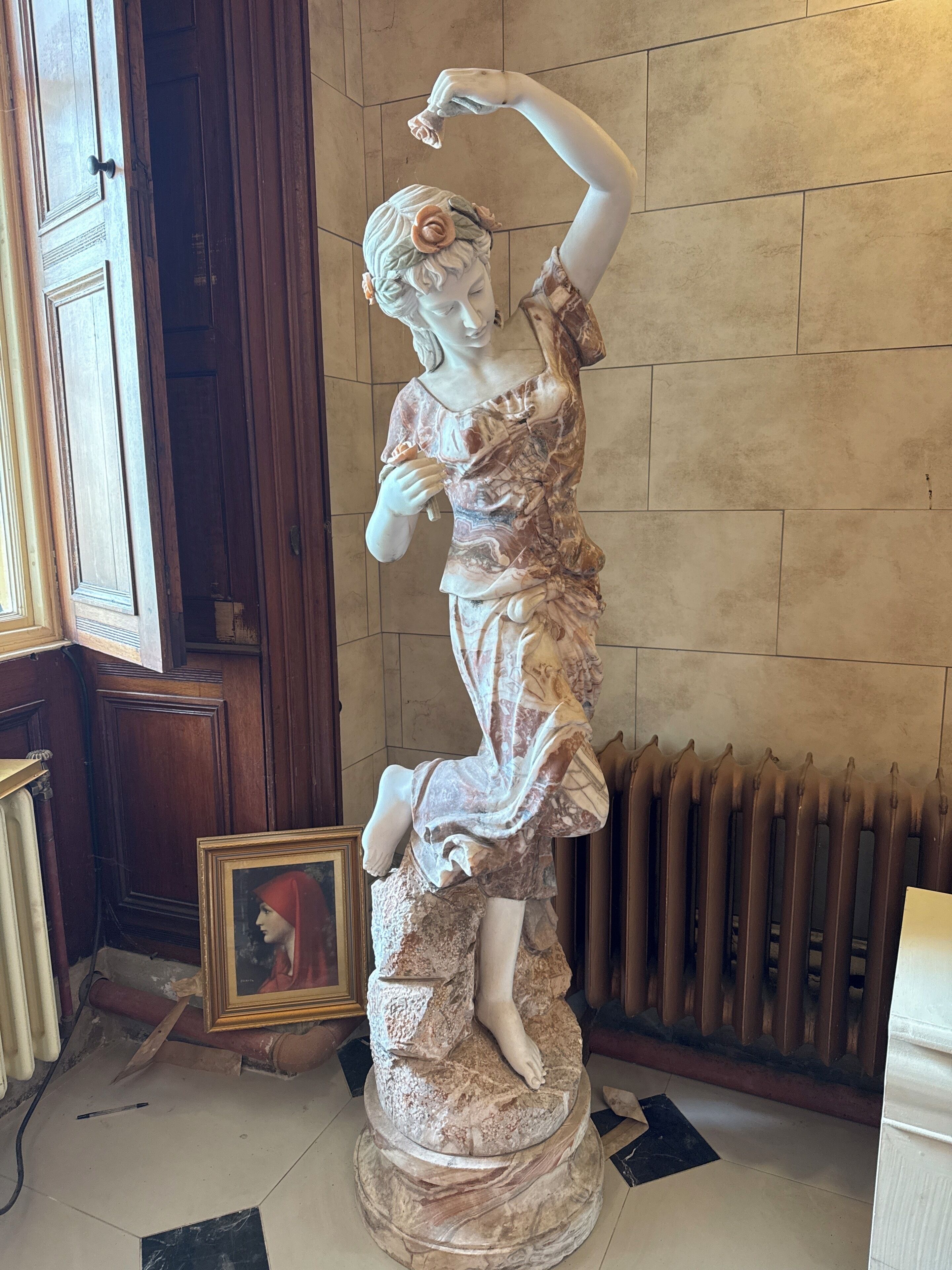 Una estatua de una mujer con vestido, de pie sobre un pedestal, con un cuadro enmarcado en el suelo a su lado.