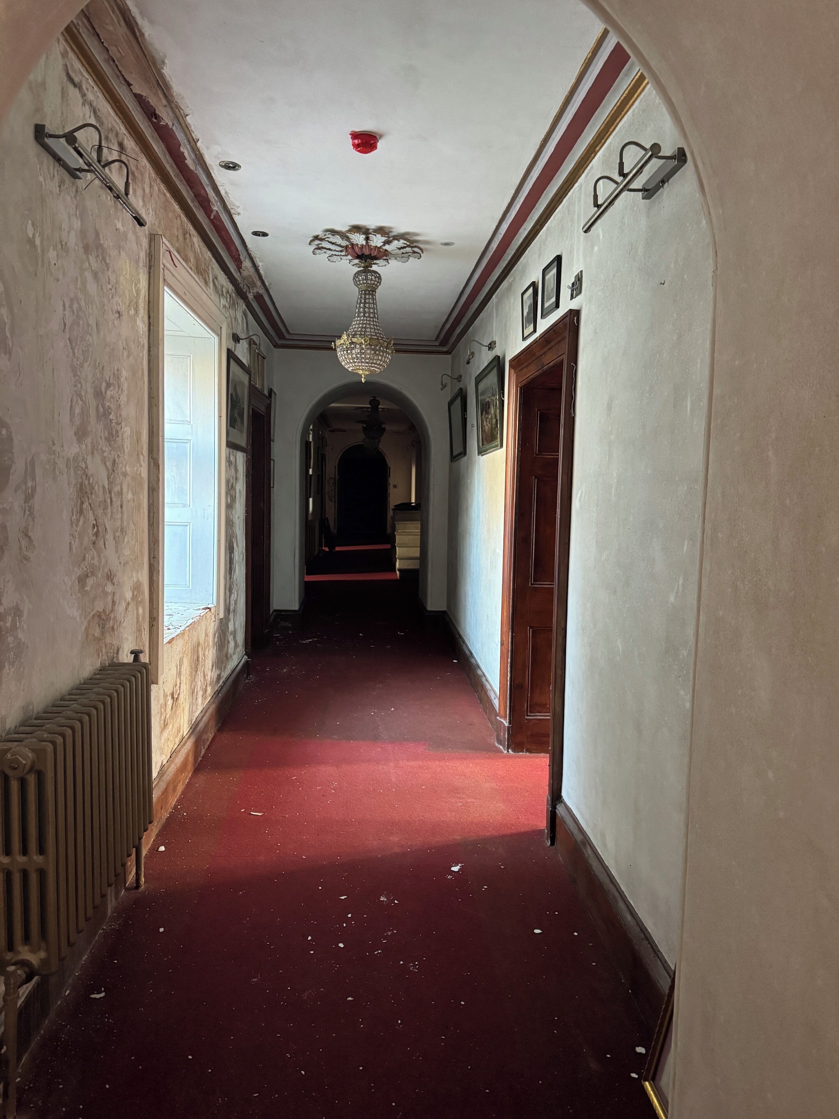 A hallway with a red carpet, a radiator, a chandelier, and a wooden door.