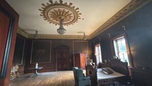Una habitación con un gran medallón decorativo en el techo, piso de madera, cama con cabecera, una silla y una ventana con cortinas.
