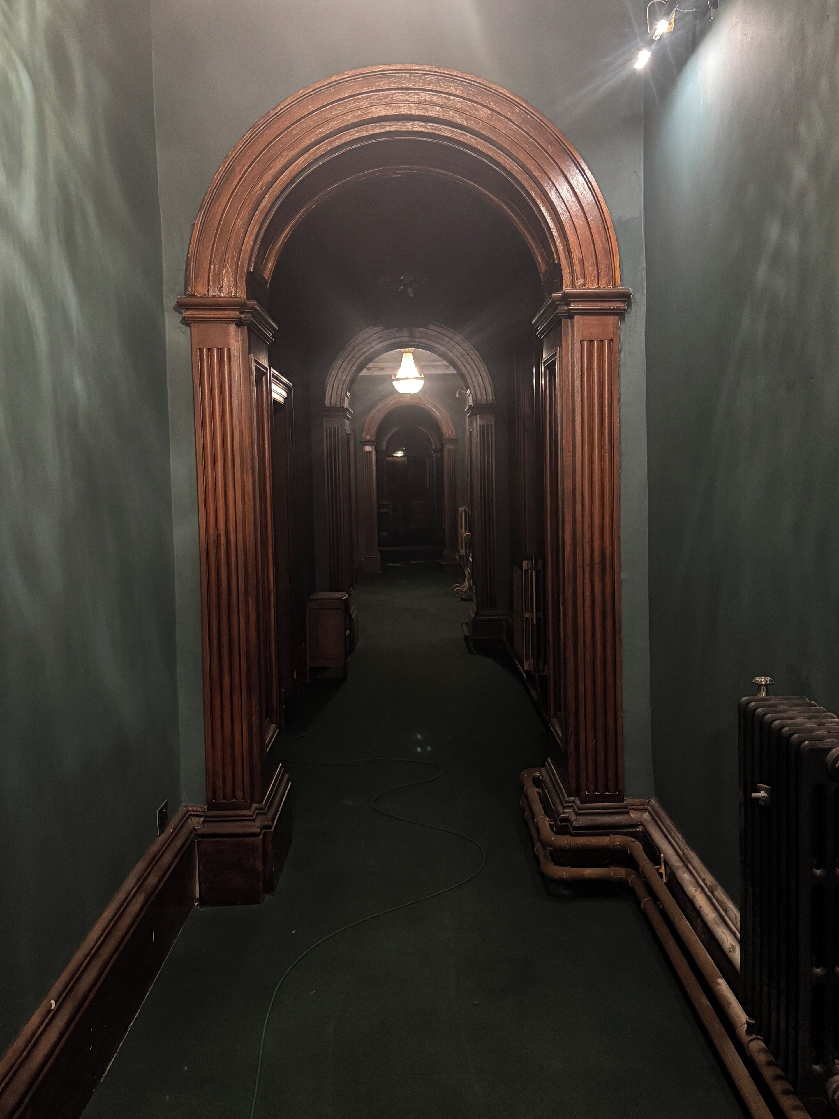 Un pasillo con columnas de madera, alfombra verde y una lámpara al final.