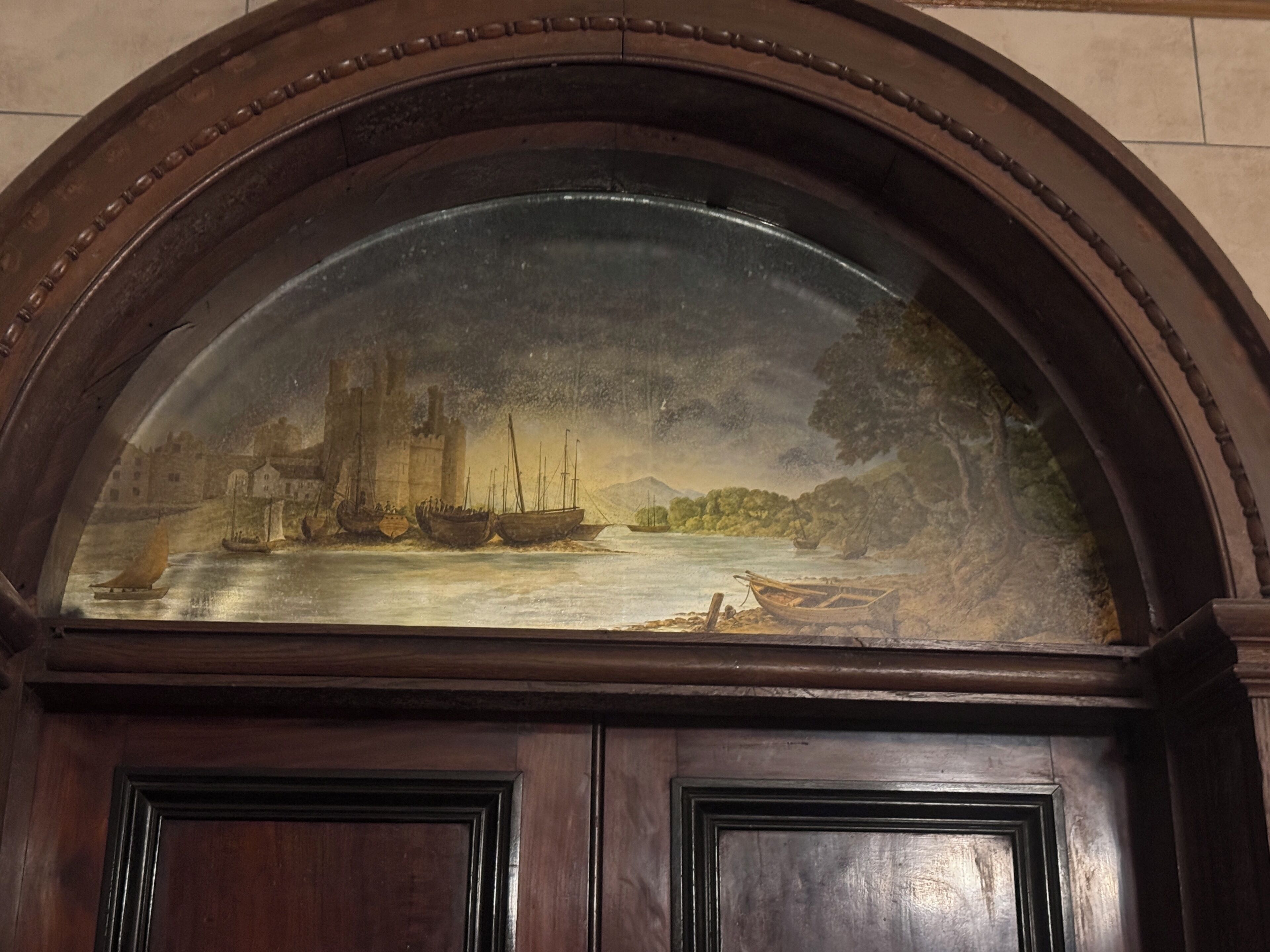 A detailed mural depicting a historical scene with ships and a castle by a river, framed in an arched wooden structure.
