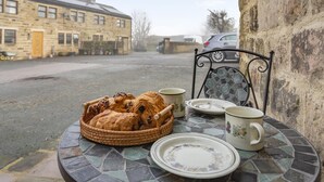 Une table avec un panier de pain, une assiette et une tasse sur une terrasse carrelée, d’où l’on aperçoit un bâtiment en pierre et une voiture garée.