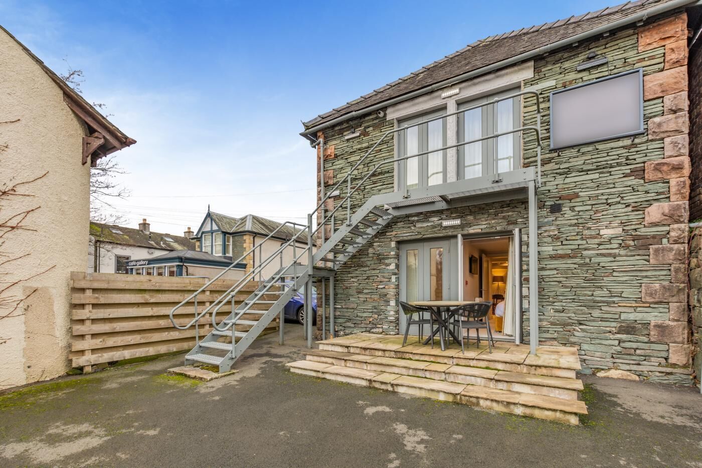 Un bâtiment en pierre avec un escalier menant à une porte, une table et des chaises à l’extérieur, et une fenêtre visible.
