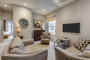 Una sala de estar moderna con sofá, sillón y una mesa de centro. Hay un televisor de pantalla plana montado en la pared, un espejo redondo y un cuadro enmarcado. La habitación tiene un gran ventanal con vistas al exterior.