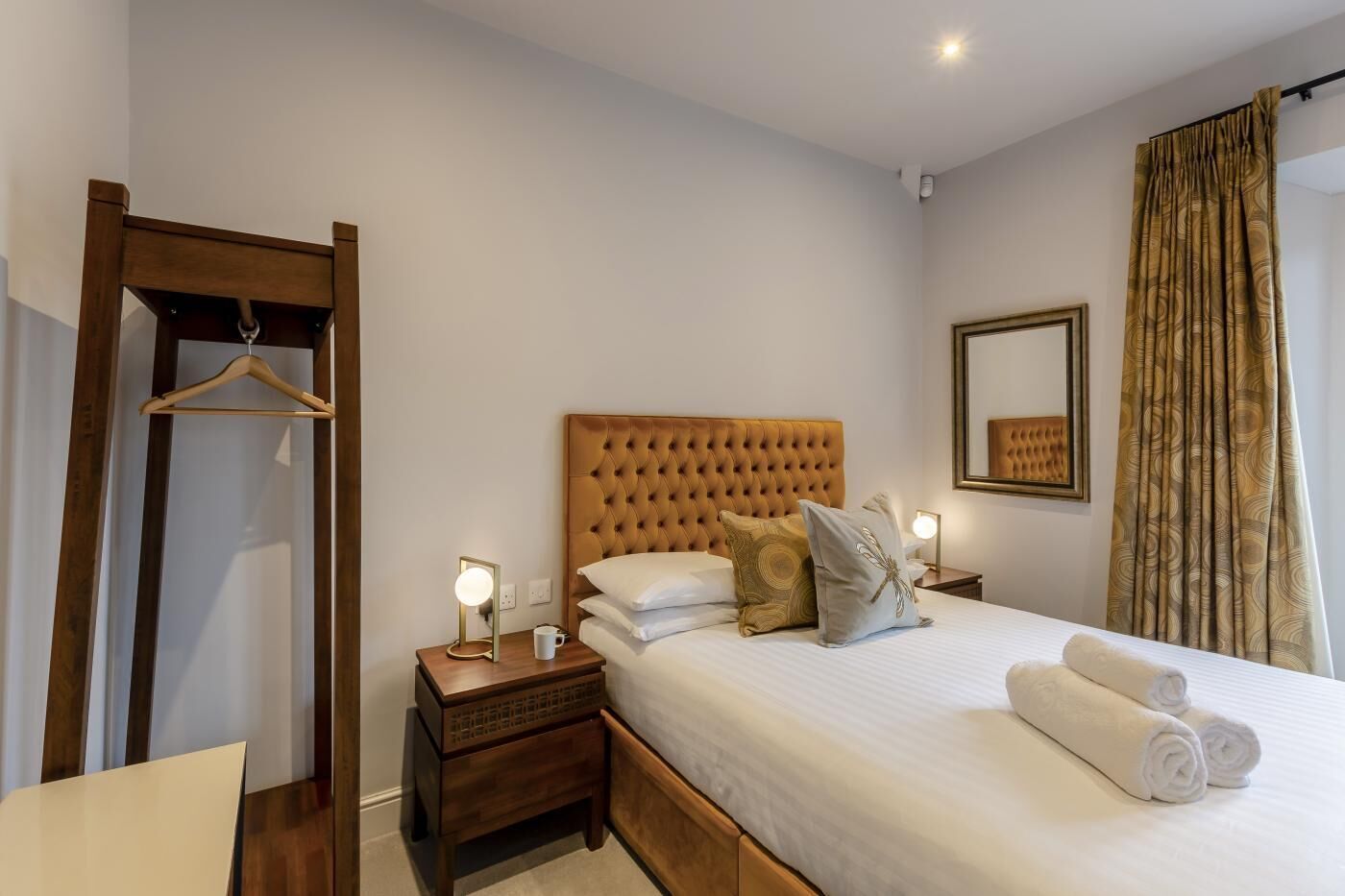 A hotel room with a large bed, a wooden nightstand, a bedside table with a lamp, a mirror, and a wardrobe.