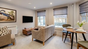 Un salon moderne comprenant un canapé, une table à manger avec des chaises, un téléviseur à écran plat et de grandes fenêtres équipées de stores.