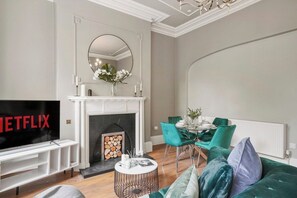 A living room with a fireplace, a TV, a dining area with teal chairs, and a green sofa. - 30% off! The Bridgeton in Wakefield Apartment (Wakefield)