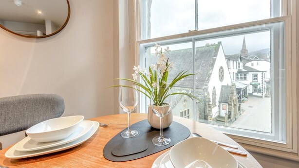 A dining table with plates, a wine glass, and a vase with flowers. A window reveals a view of buildings and a church steeple. - 1 Bed in Keswick (oc-g35025) (Keswick)