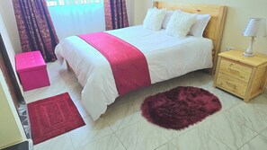 Une chambre à coucher avec un lit, une table de chevet, une tête de lit en bois, un tapis rouge et un pouf rose.