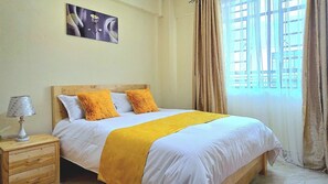 Une chambre à coucher avec un lit, une table de chevet, une lampe, un tableau au mur et une fenêtre avec des rideaux.