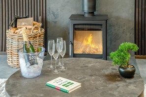 A cozy room with a fireplace, a small table with a book, a glass of champagne, and wine glasses. - 2 Bed in Plymtree (oc-m35086) (Plymtree)