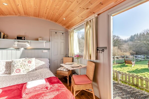 A bedroom with a bed, a small table, and a chair. There is a window with curtains, and a view of a garden outside.