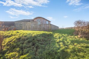 A wooden barn with a sloped roof, surrounded by a grassy field and a stone building in the background.