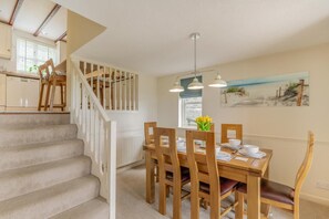 A dining area with a wooden table, chairs, and a painting of a beach scene. - 2 Bed in Looe (oc-t35020) (Looe)