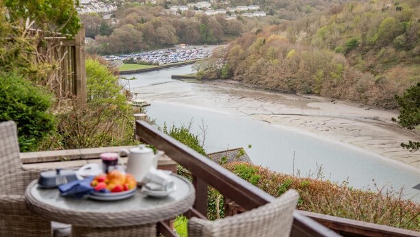 Ein Balkon mit Korbmöbeln, einem Tisch mit einer Fruchtschale und Blick auf einen Fluss mit einer Stadt im Hintergrund.