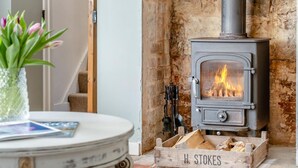 Un poêle à bois dont on peut voir les flammes, une caisse en bois portant l’inscription « H. STOKES » et contenant du bois de chauffage, ainsi qu’une table sur laquelle se trouvent une plante et un magazine.